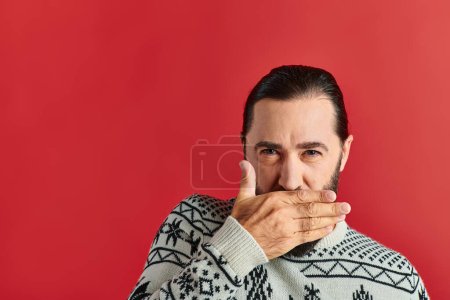 bearded man in Christmas sweater with ornament covering mouth on red backdrop, winter holidays