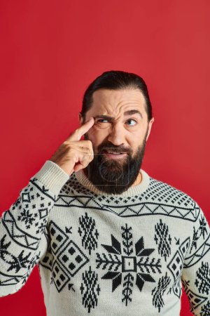 bearded man in Christmas sweater with ornament thinking and pointing at head on red background