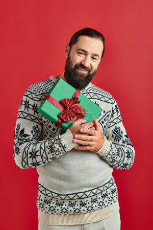 happy bearded man in winter sweater with ornament holding Christmas present on red background