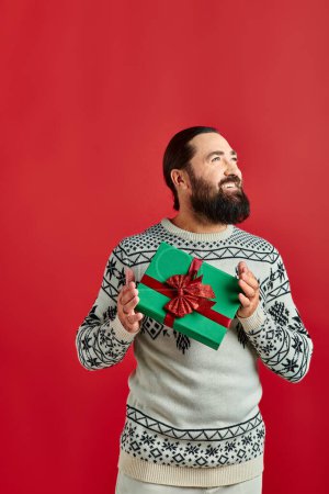 happy bearded man in winter sweater with ornament holding Christmas present on red background