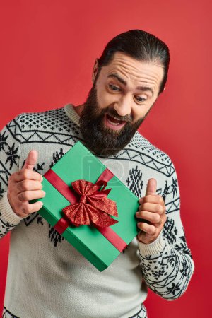 thrilled bearded man in winter sweater with ornament holding Christmas present on red background
