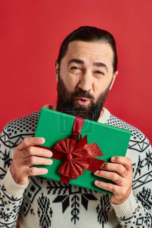 air kiss from bearded man in winter sweater with ornament holding Christmas present on red backdrop