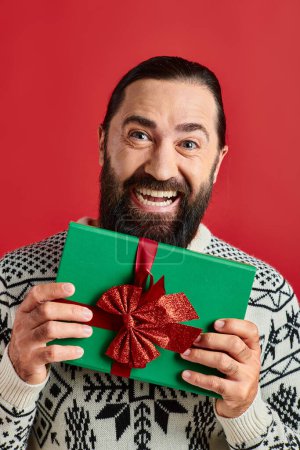 excited bearded man in winter sweater with ornament holding Christmas present on red background