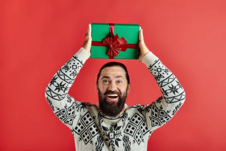 hombre barbudo excitado en suéter de invierno con adorno celebración regalo de Navidad sobre fondo rojo