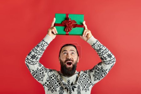 excited bearded man in winter sweater with ornament holding Christmas gift on red background