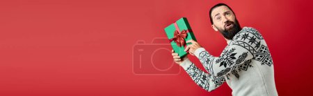 confused bearded man in winter sweater with ornament holding Christmas present on red, banner