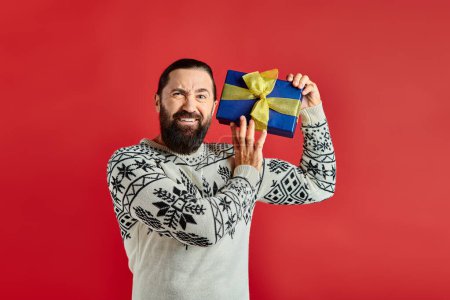 displeased bearded man in winter sweater with ornament holding Christmas present on red background