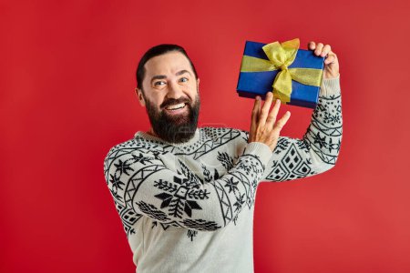 pleased bearded man in winter sweater with ornament holding Christmas present on red background