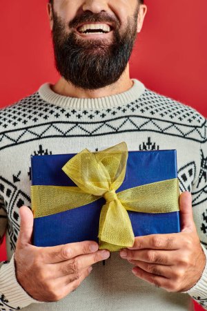 cropped happy bearded man in winter sweater with ornament holding Christmas present on red backdrop