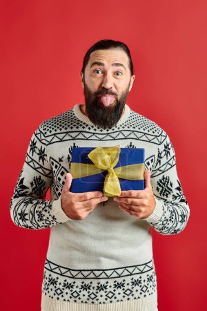 Lustiger bärtiger Mann im Winterpullover mit Ornament, der Weihnachtsgeschenk hält und Zunge auf Rot klebt
