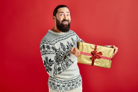 fröhlicher bärtiger Mann im Winterpullover mit Ornament hält Weihnachtsgeschenk auf rotem Hintergrund