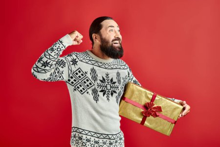 excited bearded man in winter sweater with ornament holding Christmas present on red background