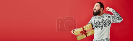 excited bearded man in winter sweater with ornament holding Christmas present on red, banner