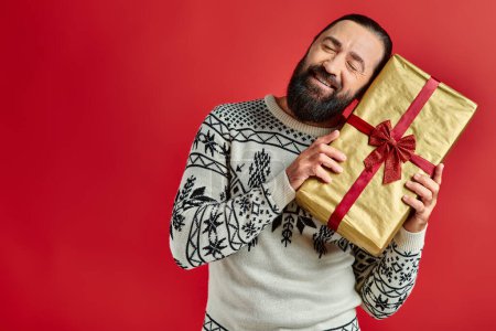 hombre barbudo alegre en suéter de invierno con adorno celebración regalo de Navidad sobre fondo rojo
