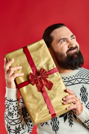 bearded displeased man in winter sweater with ornament holding Christmas present on red background