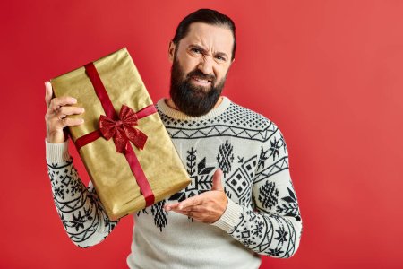 displeased bearded man in winter sweater with ornament holding Christmas present on red background