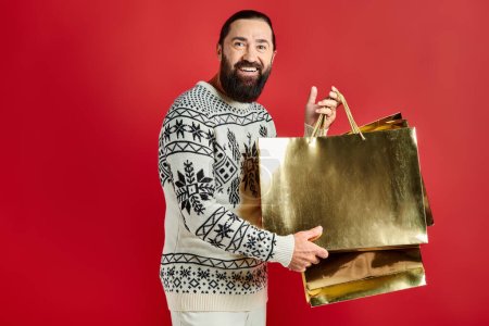 happy bearded man in sweater with ornament holding shopping bags on red backdrop, Christmas present
