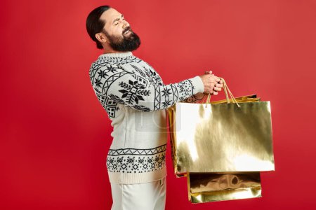 Unzufriedener bärtiger Mann im Pullover mit Ornament hält Einkaufstüten vor rotem Hintergrund, Weihnachten
