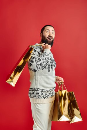 happy bearded man in sweater with ornament holding shopping bags on red backdrop, Christmas present