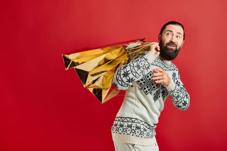 joyful bearded man in sweater with ornament holding shopping bags on red backdrop, Christmas present