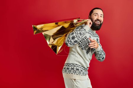 smiling bearded man in sweater with ornament holding shopping bags on red backdrop, Christmas time