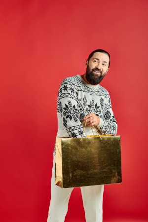 cheerful bearded man in Christmas sweater holding shopping bag on red backdrop, holiday sales