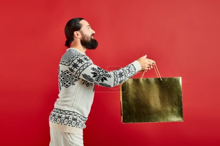 cheerful bearded man in Christmas sweater holding shopping bag on red backdrop, holiday sales