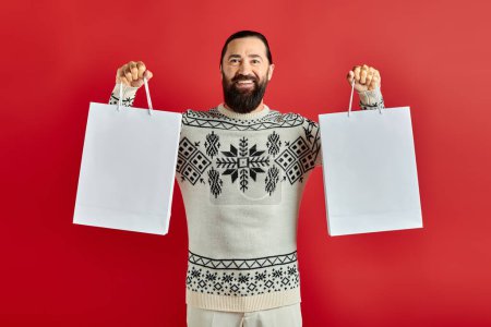 cheerful bearded man in Christmas sweater holding shopping bags on red backdrop, holiday sales