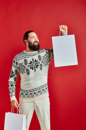 positive bearded man in Christmas sweater holding shopping bags on red backdrop, holiday sales