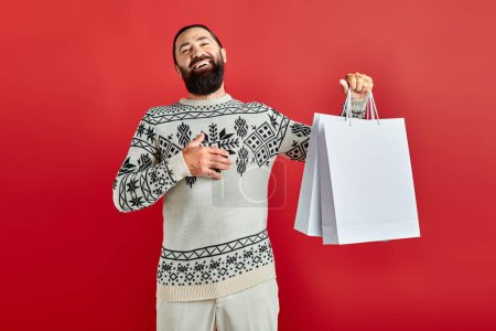 positiver bärtiger Mann im Weihnachtspulli mit Einkaufstüten vor rotem Hintergrund, Feiertagsstimmung