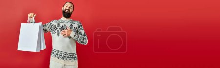 cheerful bearded man in Christmas sweater holding shopping bags on red backdrop, holiday sales