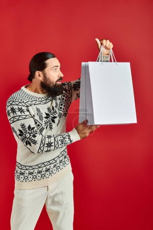 Neugieriger bärtiger Mann im Weihnachtspulli mit Einkaufstüten vor rotem Hintergrund, Feiertagsstimmung