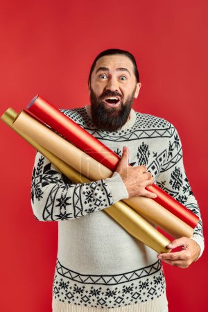 amazed bearded man in Christmas sweater holding wrapping paper on red backdrop, holiday sales