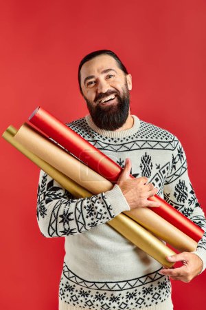 heureux homme barbu en pull de Noël tenant papier d'emballage sur fond rouge, les ventes de vacances