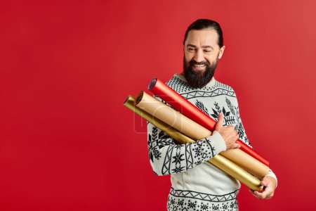 happy bearded man in Christmas sweater holding wrapping paper on red backdrop, holiday sales