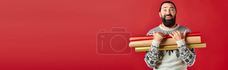 happy bearded man in Christmas sweater holding wrapping paper on red backdrop, holiday sales banner