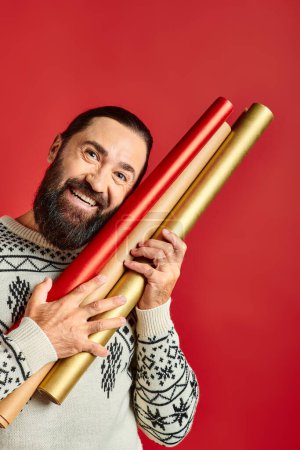 happy bearded man in winter sweater holding wrapping paper on red backdrop, Merry Christmas