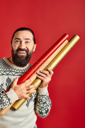 cheerful bearded man in winter sweater holding wrapping paper on red backdrop, Merry Christmas