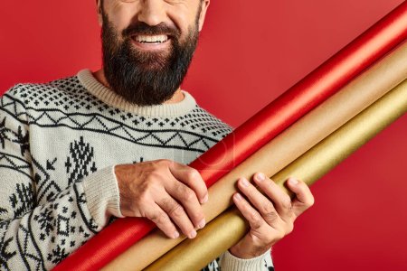 homme barbu heureux cultivé en chandail d'hiver tenant papier cadeau sur fond rouge, Joyeux Noël
