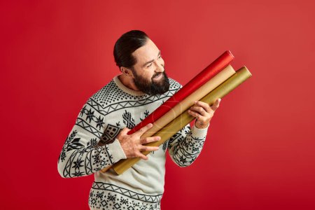 happy bearded man in winter sweater holding colorful gift paper on red backdrop, Merry Christmas