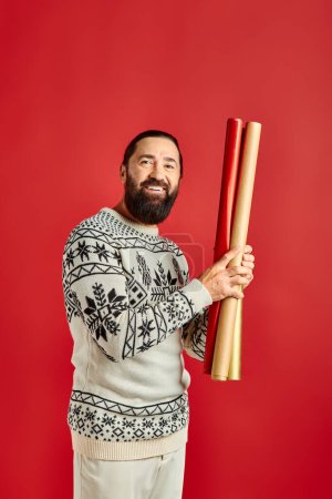 joyful bearded man in winter sweater holding colorful gift paper on red backdrop, Merry Christmas