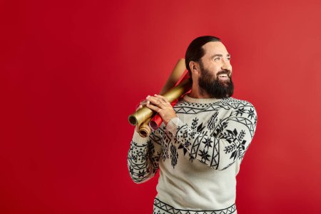joyful bearded man in winter sweater holding wrapping paper on red backdrop, Merry Christmas