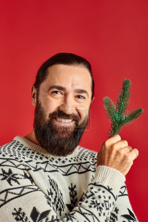 joyful bearded man in winter sweater holding branch of pine tree on red backdrop, Merry Christmas