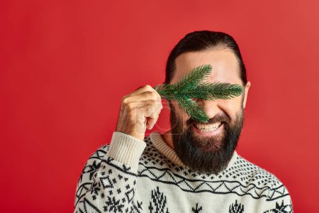joyful man in sweater covering eyes with branch of pine tree on red backdrop, Merry Christmas