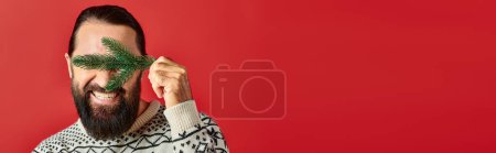 happy man in sweater covering eyes with branch of pine tree on red backdrop, Merry Christmas banner