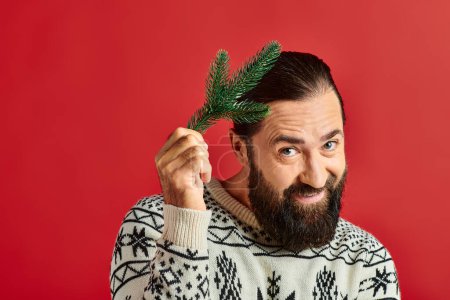 happy bearded man in winter sweater holding branch of pine tree on red backdrop, Merry Christmas