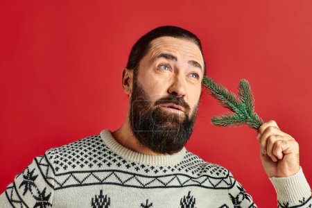 pensive bearded man in winter sweater holding branch of pine tree on red backdrop, Merry Christmas