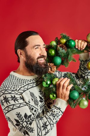 happy bearded man in winter sweater holding decorated wreath with baubles on red backdrop, Christmas