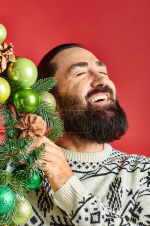 cheerful bearded man in winter sweater holding Christmas wreath with baubles on red backdrop