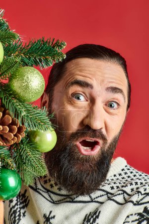 positive bearded man in winter sweater holding Christmas wreath with baubles on red backdrop